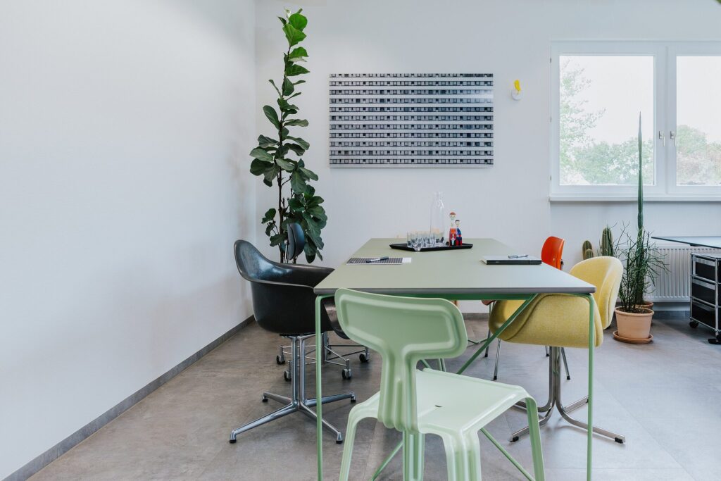 Modernes Büro mit Tisch, bunten Stühlen, Pflanzen und einem Kunstwerk an der Wand. Helle, einladende Atmosphäre.