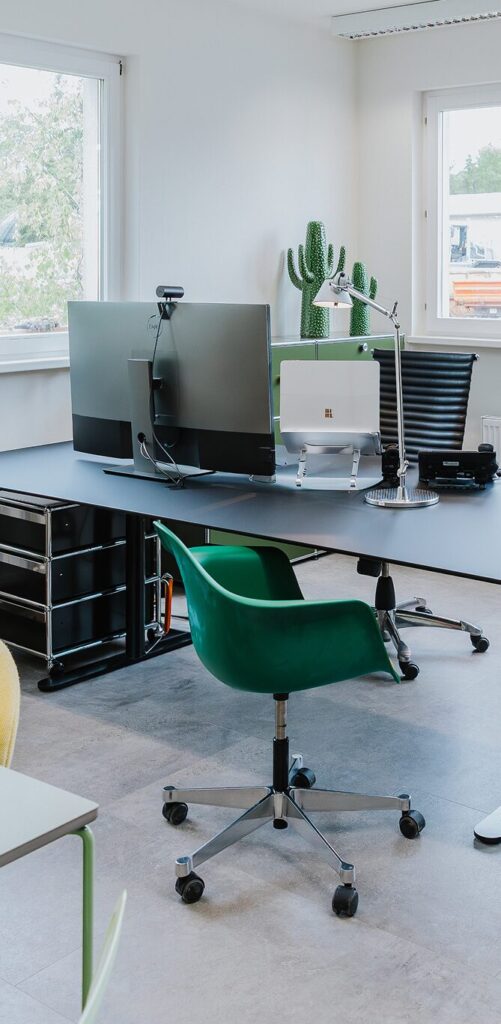 Heller Arbeitsplatz mit zwei großen Monitoren, einem grünen Stuhl und einem Kaktus in der Ecke.
