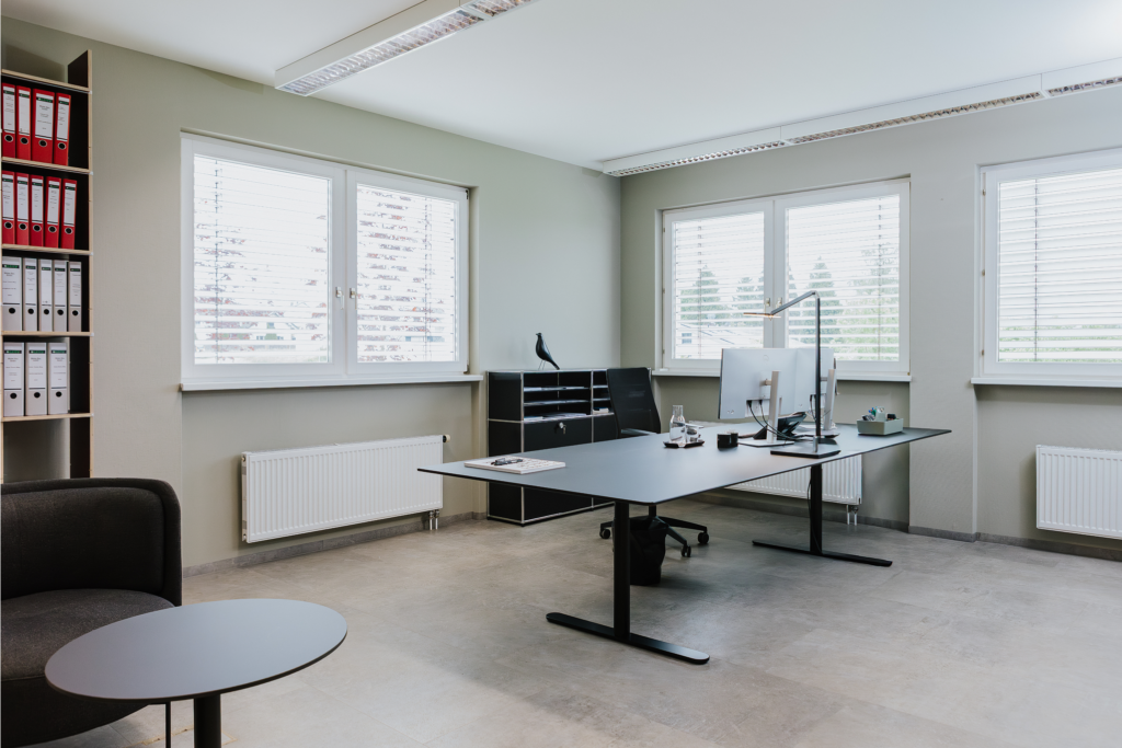 Modernes Büro mit Schreibtisch, Computer, Stuhl, Regalen voller Akten und viel natürlichem Licht durch Fenster.